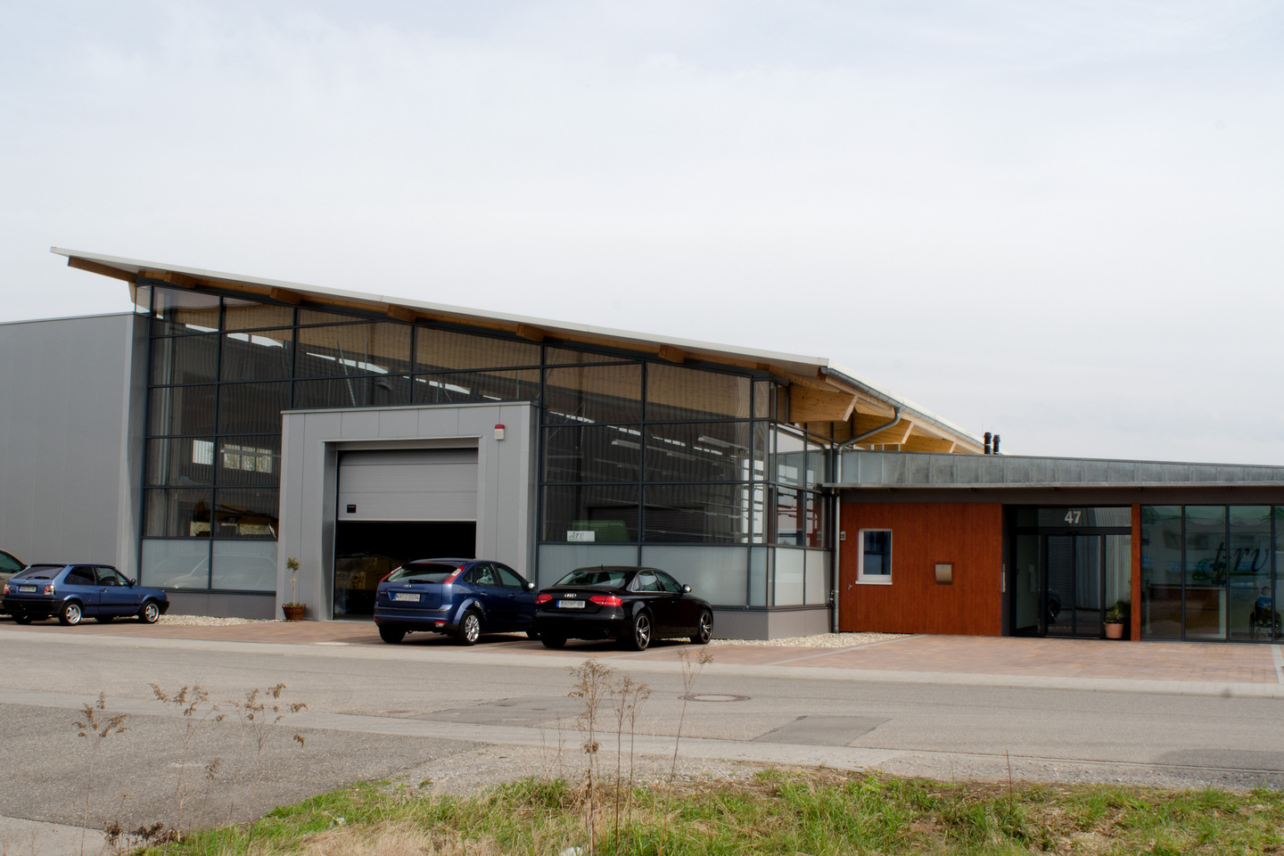 Ein modernes Gebäude mit großen Fenstern und Garagentoren, vor dem mehrere Autos geparkt sind.